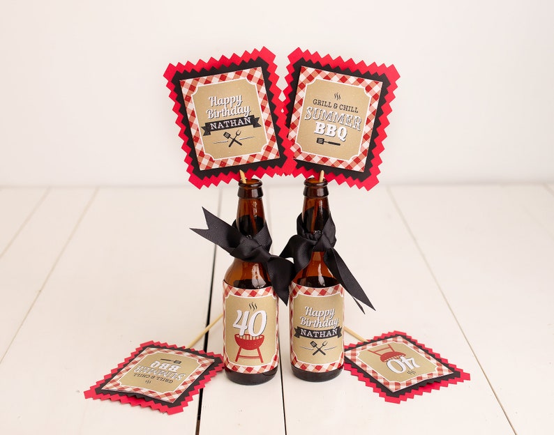 Puede incluir: Dos botellas de cerveza marrones decoradas con cintas negras y etiquetas personalizadas. Las etiquetas dicen "Happy Birthday Nathan" y "Grill & Chill Summer BBQ". Adornos a cuadros rojos y negros en palitos de madera realzan el tema de la barbacoa.
