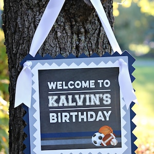 May include: A blue and gray welcome sign with a white ribbon hanging from a tree branch. The sign says "Welcome to Kalvin's Birthday" with a basketball, football, and baseball graphic.