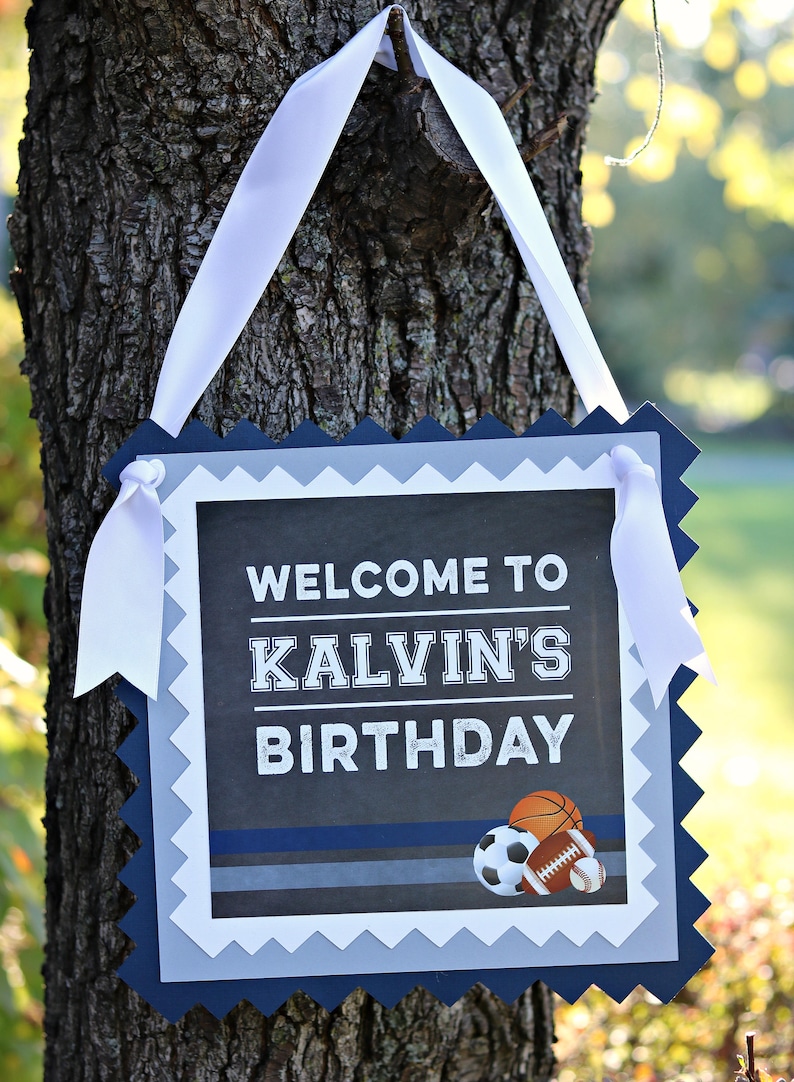 May include: A blue and gray welcome sign with a white ribbon hanging from a tree branch. The sign says "Welcome to Kalvin's Birthday" with a basketball, football, and baseball graphic.