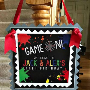 May include: A black and white sign with a gray scalloped border. The sign says "Game On!" with a target symbol. Below the text, it says "Welcome to Jack & Alex's 11th Birthday" with colorful paint splatters in the background.