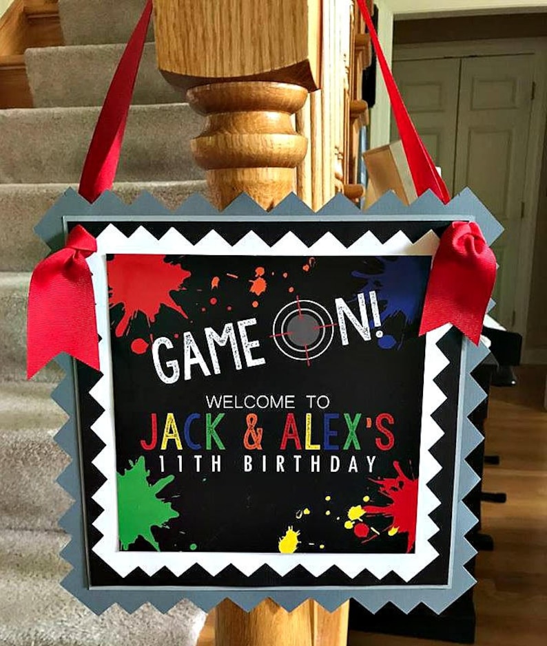 May include: A black and white sign with a gray scalloped border. The sign says "Game On!" with a target symbol. Below the text, it says "Welcome to Jack & Alex's 11th Birthday" with colorful paint splatters in the background.