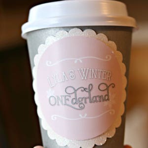 May include: A gray paper coffee cup with a white lid. A pink and white label with the text "Lyla's Winter ONEderland" is affixed to the cup. The label has a scalloped edge and snowflake accents.