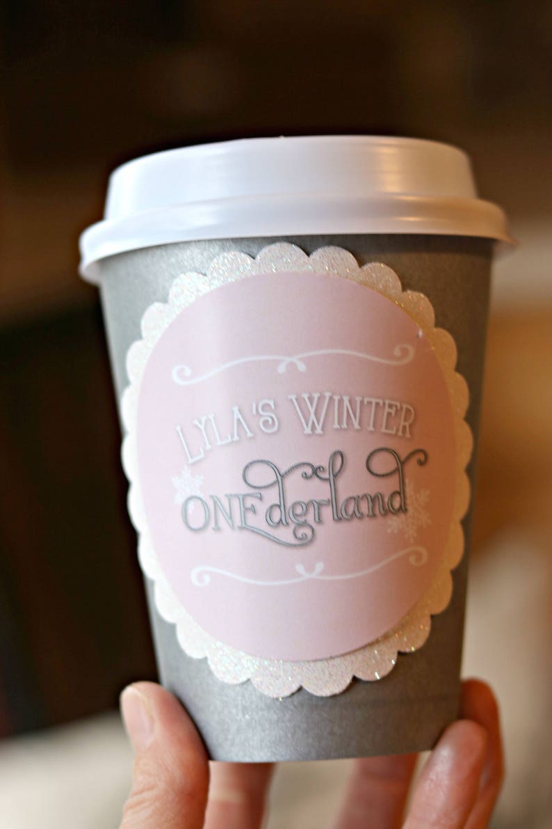 May include: A gray paper coffee cup with a white lid. A pink and white label with the text "Lyla's Winter ONEderland" is affixed to the cup. The label has a scalloped edge and snowflake accents.