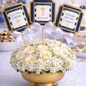 May include: A floral arrangement in a gold-colored bowl, featuring white roses and small white flowers. Three navy blue and gold-trimmed signs with white bows are placed in the arrangement. The signs have text that reads "God Bless Taylor", "May 8, 2019", and "Taylor's First Holy Communion".