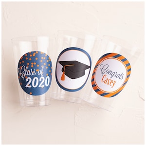 May include: Three clear plastic cups with graduation themed stickers. The first cup has a blue and orange sticker with the text "Class of 2020". The second cup has a blue and orange sticker with a black graduation cap. The third cup has a blue and orange striped sticker with the text "Congrats Casey".