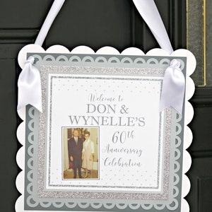 May include: A white sign with a scalloped edge and a gray border. The sign has a photo of an older couple, the text "Welcome to Don & Wynnelle's 60th Anniversary Celebration", and a silver glitter background.