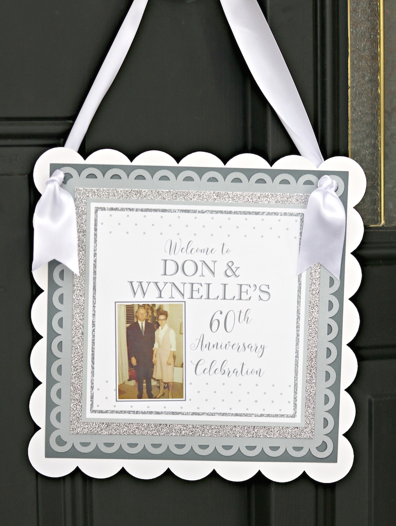 May include: A white sign with a scalloped edge and a gray border. The sign has a photo of an older couple, the text "Welcome to Don & Wynnelle's 60th Anniversary Celebration", and a silver glitter background.