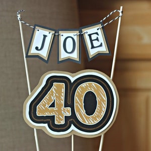 May include: A cake topper with a black and gold banner that says "JOE" and a large black and gold number "40" for a birthday celebration.