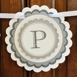 May include: A white banner with a scalloped edge and a silver glitter circle with the letter "P" in the center. The banner is strung with white ribbon.