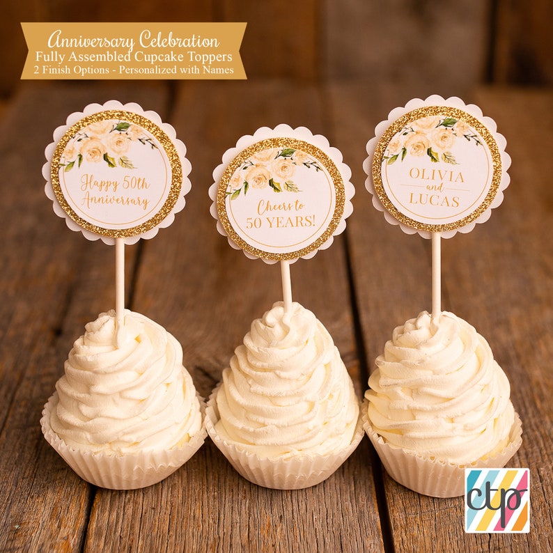 K&ouml;nnte beinhalten: Drei wei&szlig;e Cupcakes mit goldenen Glitzer-Cupcake-Toppern. Die Topper zeigen wei&szlig;e Rosen und goldenen Glitzer mit dem Text "Happy 50th Anniversary", "Cheers to 50 Years!" und "Olivia and Lucas".
