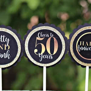 May include: Three black and gold cupcake toppers with scalloped edges. The first topper says "Betty AND Frank". The second topper says "Cheers to 50 Years". The third topper says "Happy Anniversary".