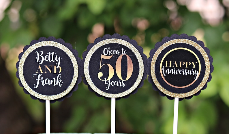 May include: Three black and gold cupcake toppers with scalloped edges. The first topper says "Betty AND Frank". The second topper says "Cheers to 50 Years". The third topper says "Happy Anniversary".