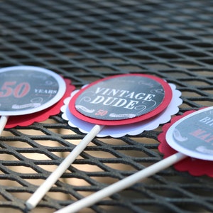 May include: Three round cupcake toppers with scalloped edges. The toppers are black and white with red accents. The text on the toppers reads "Aged 50 Years", "Vintage Dude", and "Happy Birthday Matt".