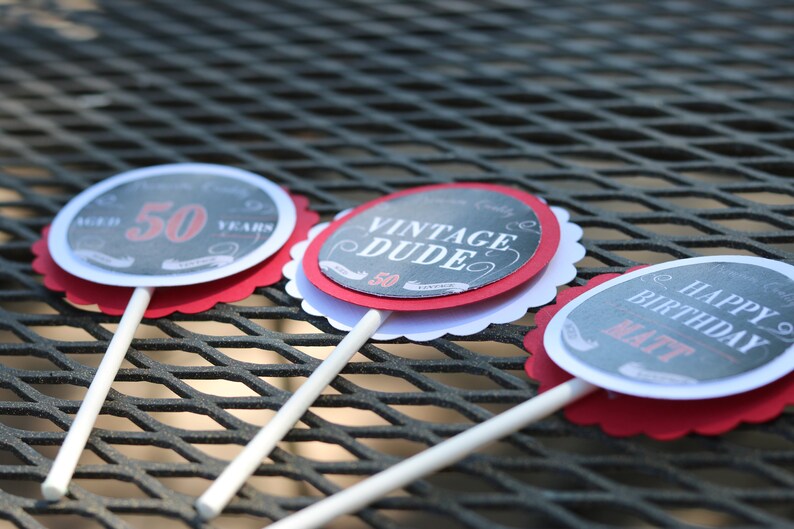 May include: Three round cupcake toppers with scalloped edges. The toppers are black and white with red accents. The text on the toppers reads "Aged 50 Years", "Vintage Dude", and "Happy Birthday Matt".