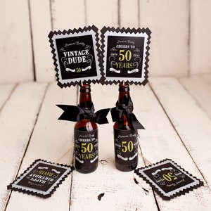 以下が含まれることがあります： 黒と金のラベルが付いた茶色のガラス瓶2本。ラベルには「Cheers to 50 Years!」と「Vintage Dude 50」と書かれている。瓶は黒いリボンで飾られており、白い木製の表面の上に置かれている。黒と銀のサインが4つあり、金色の文字で「Cheers to 50 Years!」と「Vintage Dude 50」と書かれている。