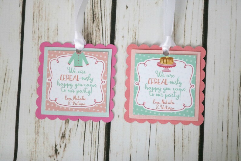 May include: Two pink and green favor tags with scalloped edges. The tags feature a cereal bowl with a spoon and a cereal box with a milk carton. The text on the tags reads "We are CEREAL-ously happy you came to our party! Love, Natalie & Victoria".