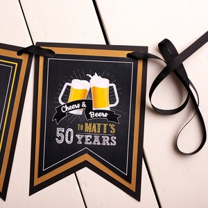 Op de afbeelding: Een zwarte banner met gouden en witte accenten, met de tekst "Cheers & Beers to Matt's 50 Years" en illustraties van bierpullen. De banner is geregen met een zwart lint, perfect voor een mijlpaal verjaardag.