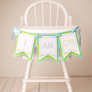 May include: A blue and green banner with the words "I am one" on it. The banner is hanging from a white high chair.