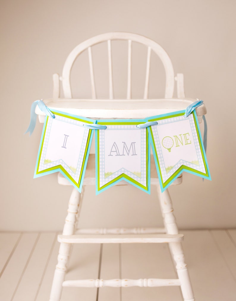 May include: A blue and green banner with the words "I am one" on it. The banner is hanging from a white high chair.