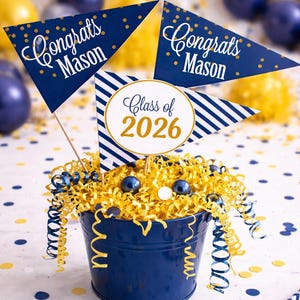 May include: A navy blue bucket filled with yellow shredded paper, blue and white confetti, and decorative flags. The flags read "Congrats Mason" and "Class of 2026". The arrangement is decorated with blue and yellow streamers and balloons, perfect for a graduation celebration.