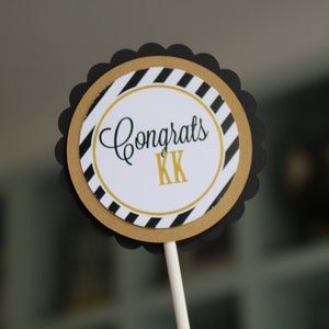 May include: Black and gold striped cupcake topper with the text "Congrats KK" in black script.