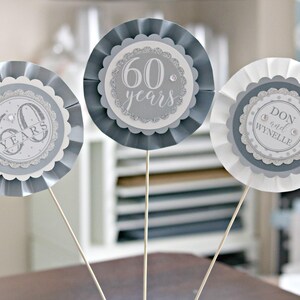 May include: Three silver and white paper rosettes with a scalloped edge. The centre of each rosette features a silver circle with a white design. One rosette says "60 years", another says "60 years", and the third says "Don and Wynnelie".