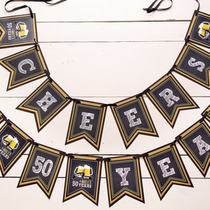 Op de afbeelding: Een zwart en goudkleurig spandoek met de tekst "Cheers to 50 Years". Elke vlag heeft een gouden rand en witte tekst. Sommige vlaggen hebben een biermok-afbeelding. De banner is geregen op een zwart lint.