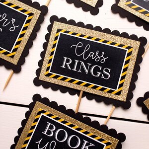 May include: Black and gold graduation party decorations with scalloped edges. The signs read "Class Rings", "Book Worms", and "Seniors".