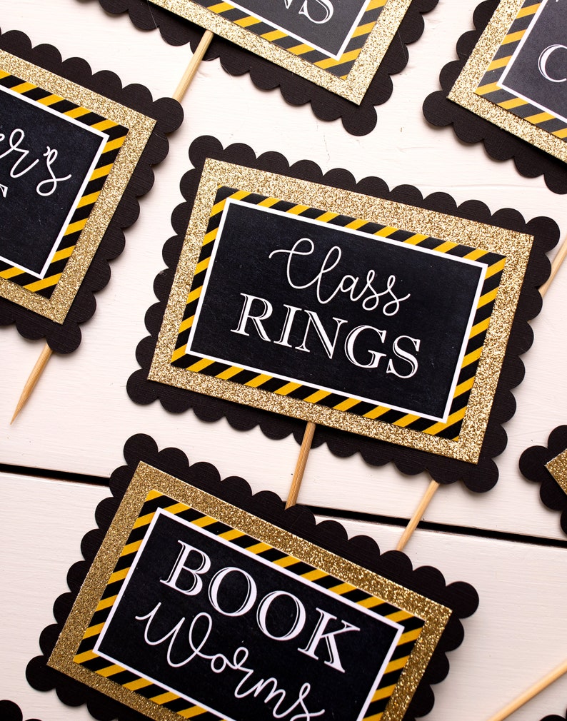 May include: Black and gold graduation party decorations with scalloped edges. The signs read "Class Rings", "Book Worms", and "Seniors".