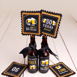 May include: Two small beer bottles with black ribbons and labels that say "Happy Birthday Matt" and "Cheers & Beers to Matt's 50 Years". There are also four black and gold party decorations with the numbers "50" and the text "50 Years Loved".