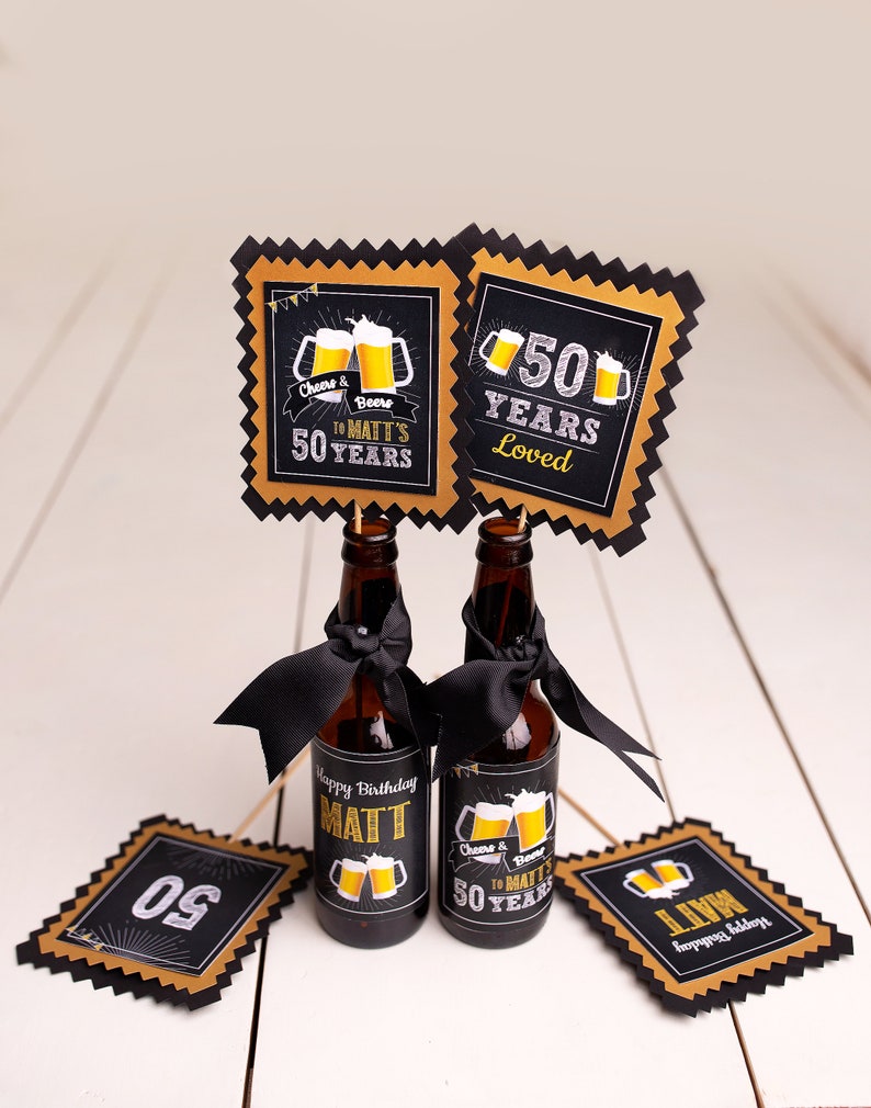 May include: Two small beer bottles with black ribbons and labels that say "Happy Birthday Matt" and "Cheers & Beers to Matt's 50 Years". There are also four black and gold party decorations with the numbers "50" and the text "50 Years Loved".