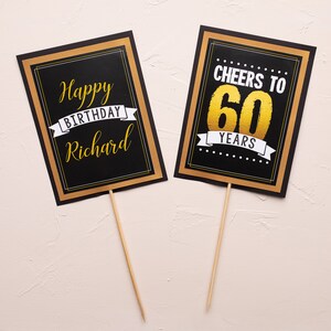 May include: Two black and gold signs with white text. The first sign says "Happy Birthday Richard" with a gold ribbon. The second sign says "Cheers to 60 Years" with a gold ribbon.
