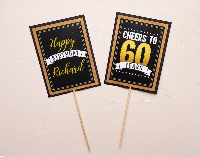 May include: Two black and gold signs with white text. The first sign says "Happy Birthday Richard" with a gold ribbon. The second sign says "Cheers to 60 Years" with a gold ribbon.