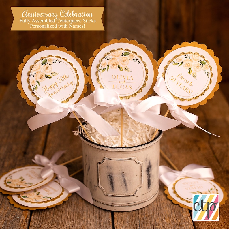 K&ouml;nnte beinhalten: Goldene und wei&szlig;e runde Tischdekorationssticks mit Blumendesigns und Text, der "Happy 50th Anniversary", "Olivia and Lucas" und "Cheers to 50 Years!" lautet.