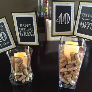 May include: A table setting for a 40th birthday party with black and gold decorations. There are four signs with the text "Cheers to 40 Years", "Happy Birthday Greg", "40" and "Vintage 1975". Two glass vases filled with wine corks and candles are on the table.