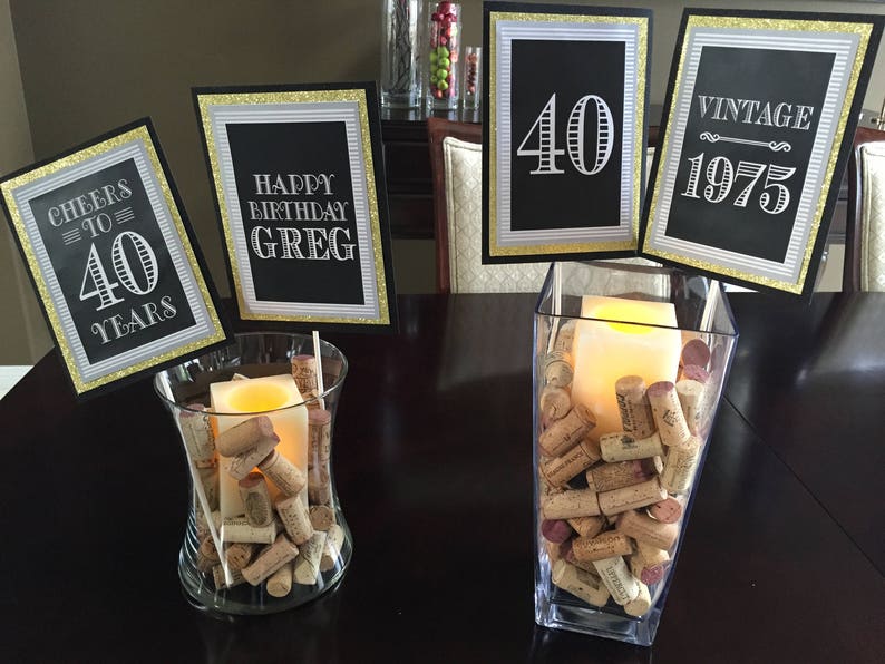 May include: A table setting for a 40th birthday party with black and gold decorations. There are four signs with the text "Cheers to 40 Years", "Happy Birthday Greg", "40" and "Vintage 1975". Two glass vases filled with wine corks and candles are on the table.