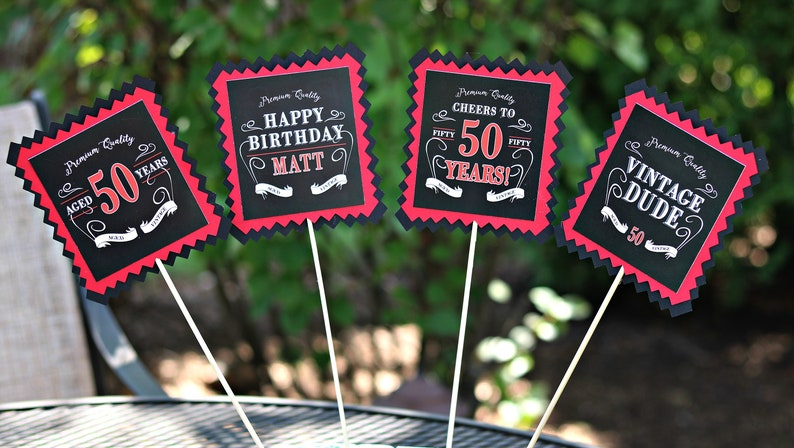 May include: Four black and red party signs with white text. The signs say "Aged 50 Years", "Happy Birthday Matt", "Cheers to 50 Years!" and "Vintage Dude 50".