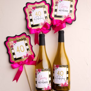 Puede incluir: Tres botellas de vino con etiquetas que dicen "Cheers to 40 Years" y "40 & Fabulous" con rayas blancas y negras, purpurina dorada y flores rosas. Tambi&eacute;n hay dos carteles rosas y dorados que dicen "40 & Fabulous" y "Cheers to 40 Years".