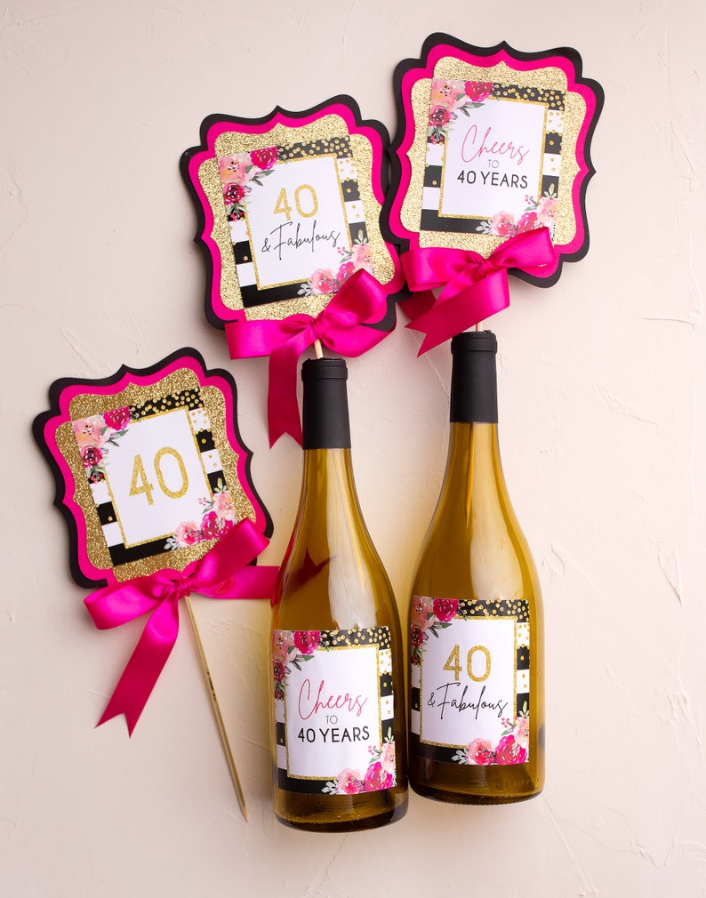 Puede incluir: Tres botellas de vino con etiquetas que dicen "Cheers to 40 Years" y "40 & Fabulous" con rayas blancas y negras, purpurina dorada y flores rosas. Tambi&eacute;n hay dos carteles rosas y dorados que dicen "40 & Fabulous" y "Cheers to 40 Years".