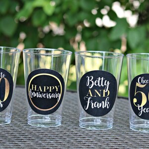 May include: Four clear plastic cups with black and gold round stickers. The stickers say "Cheers to 50 Years", "Happy Anniversary", "Betty and Frank", and "Cheers to 50 Years".