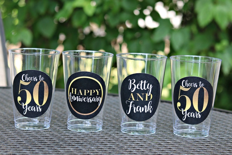 May include: Four clear plastic cups with black and gold round stickers. The stickers say "Cheers to 50 Years", "Happy Anniversary", "Betty and Frank", and "Cheers to 50 Years".