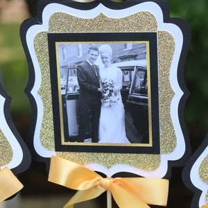 May include: Black and gold glitter frame with a white border, featuring a black and white photo of a couple in front of a car. The frame is attached to a yellow ribbon and a wooden stick.