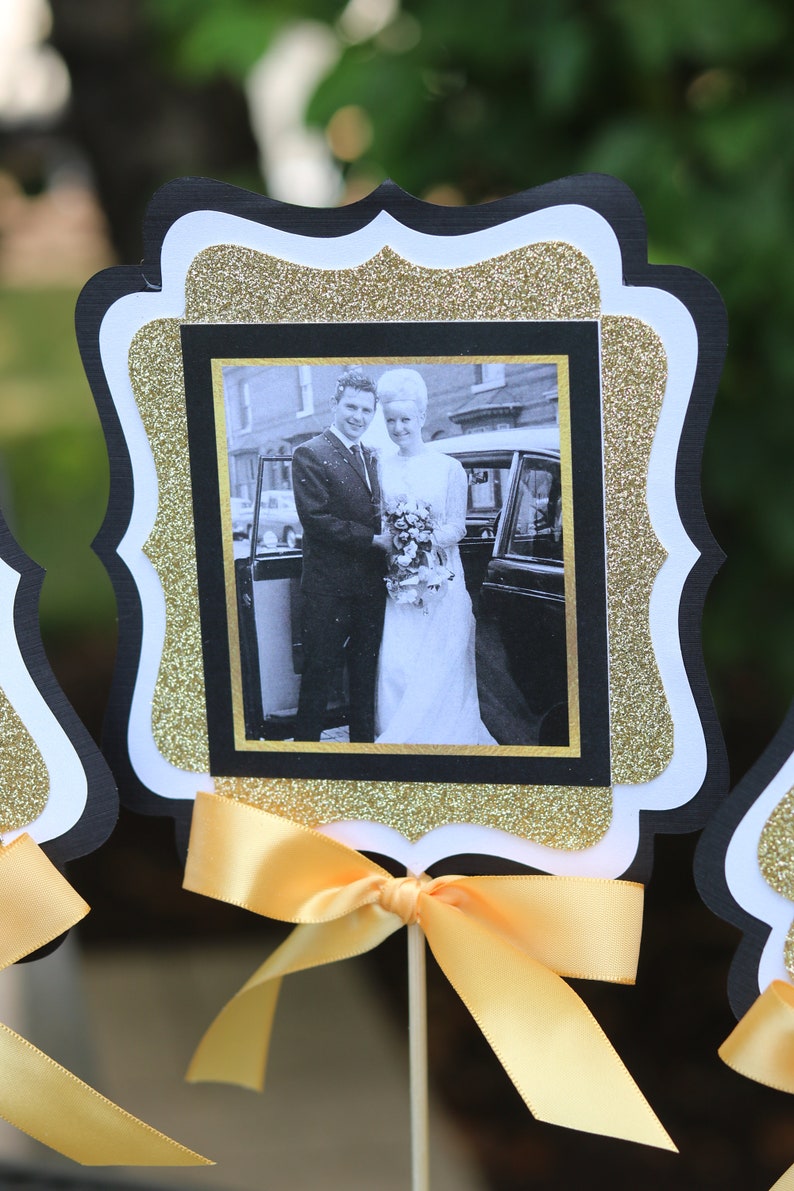 May include: Black and gold glitter frame with a white border, featuring a black and white photo of a couple in front of a car. The frame is attached to a yellow ribbon and a wooden stick.