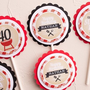 Peut inclure: Plusieurs d&eacute;corations de cupcakes avec des bords festonn&eacute;s. Les d&eacute;corations pr&eacute;sentent un motif &agrave; carreaux rouges et blancs, avec le texte "Happy Birthday Nathan", "Grill & Chill Summer BBQ", et le chiffre "40" avec une illustration de grill. Les d&eacute;corations sont sur des b&acirc;tonnets en bois.