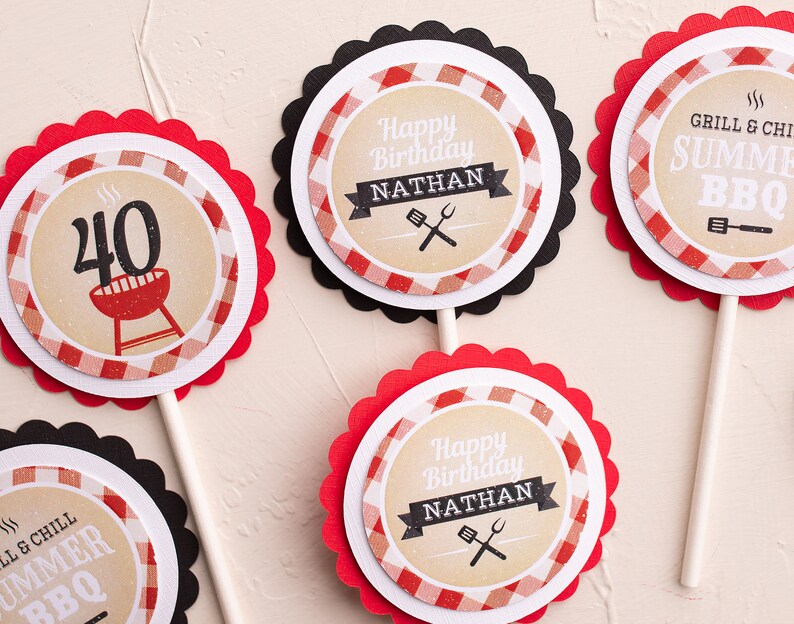 Peut inclure: Plusieurs d&eacute;corations de cupcakes avec des bords festonn&eacute;s. Les d&eacute;corations pr&eacute;sentent un motif &agrave; carreaux rouges et blancs, avec le texte "Happy Birthday Nathan", "Grill & Chill Summer BBQ", et le chiffre "40" avec une illustration de grill. Les d&eacute;corations sont sur des b&acirc;tonnets en bois.