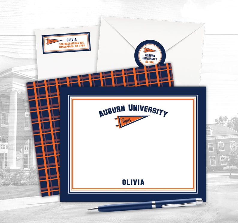 May include: A stationery set with Auburn University branding. The set includes a navy and orange plaid card, an envelope, a sticker, and a personalized card with the name "Olivia". A blue pen is also visible.