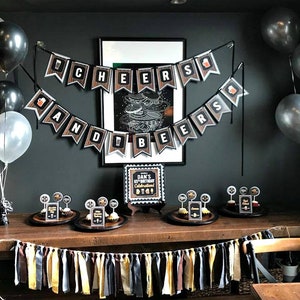 May include: A party table decorated with black, gold, and white balloons, a banner that reads "Cheers and Beers", and cupcakes with black and gold toppers. The table is set with a black and gold sign that reads "Dan's 65th Birthday Celebration Big 6-5".