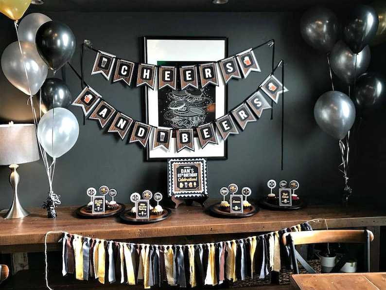 May include: A party table decorated with black, gold, and white balloons, a banner that reads "Cheers and Beers", and cupcakes with black and gold toppers. The table is set with a black and gold sign that reads "Dan's 65th Birthday Celebration Big 6-5".