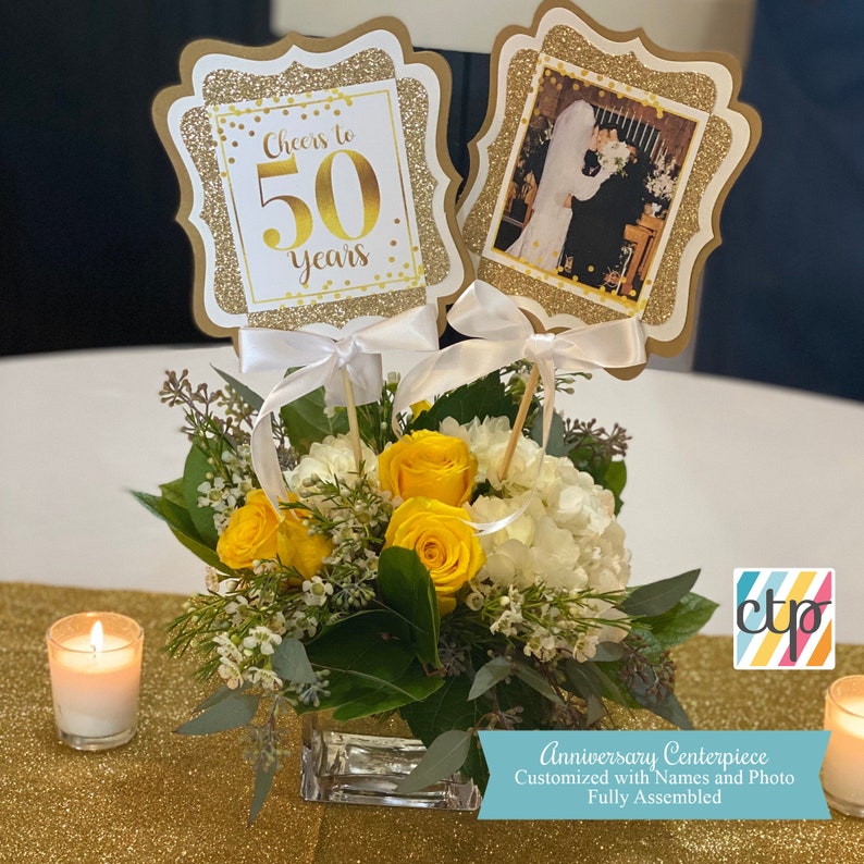 May include: Gold and white centerpiece with a photo of a couple and a sign that says "Cheers to 50 years".