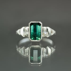May include: A silver ring featuring a rectangular, emerald-cut green gemstone as the centerpiece. The ring has two triangular, clear gemstones on each side of the main stone. The ring is set against a dark gray background, with a reflection visible below.